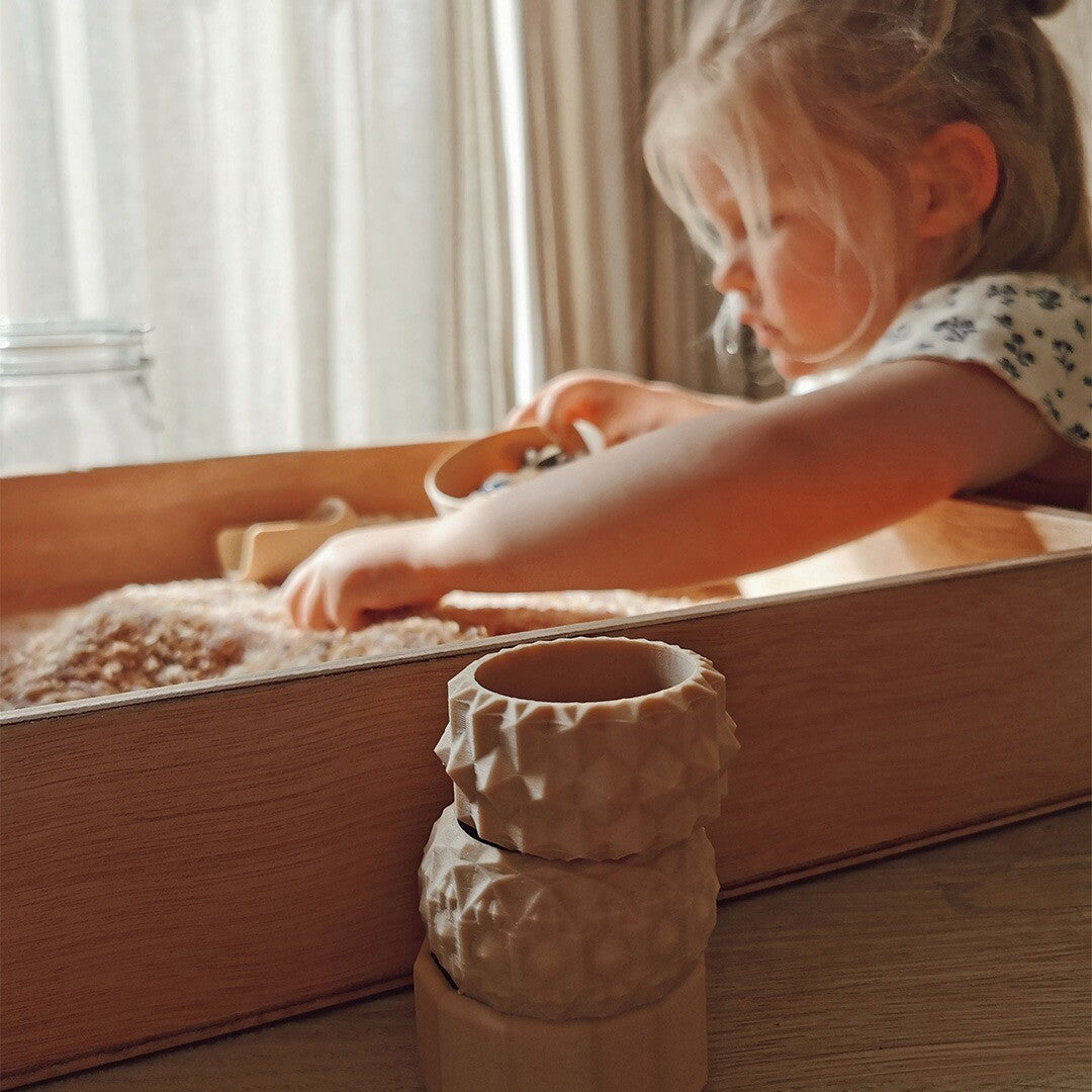 Een kind speelt met grennn mini speelbakjes in een houten speelbak met zand, met twee gestapelde bakjes op de voorgrond.