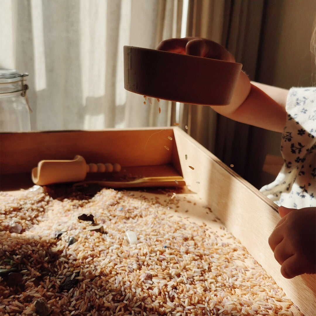 Een kinderhand laat rijst uit de grennn mini zeef vallen in een houten bak met sensorisch speelgoed.