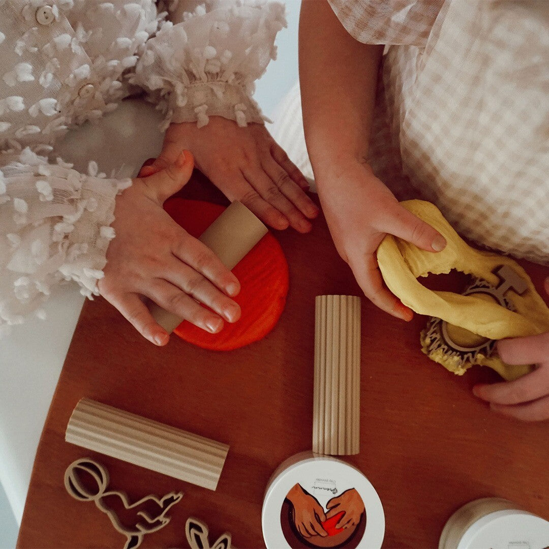 Twee kinderen spelen met rode en gele klei en houten kleirollers op een houten tafel.