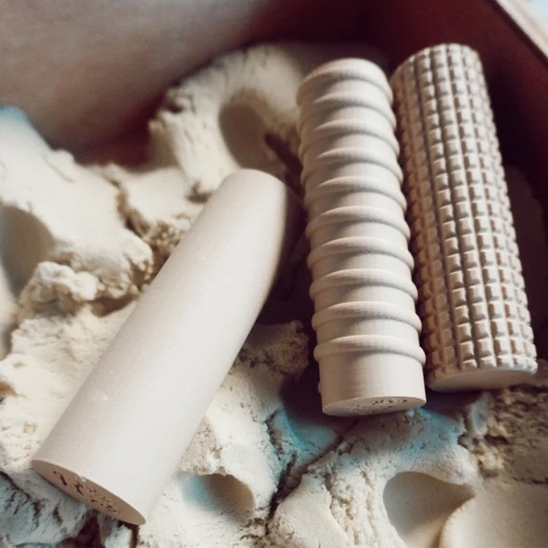 Drie verschillende houten kleirollers met patronen liggen in beige speelzand.