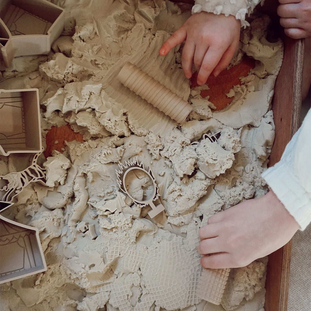 Twee kinderhanden spelen met grennn houten rollers en uitsteekvormen in beige speelzand.