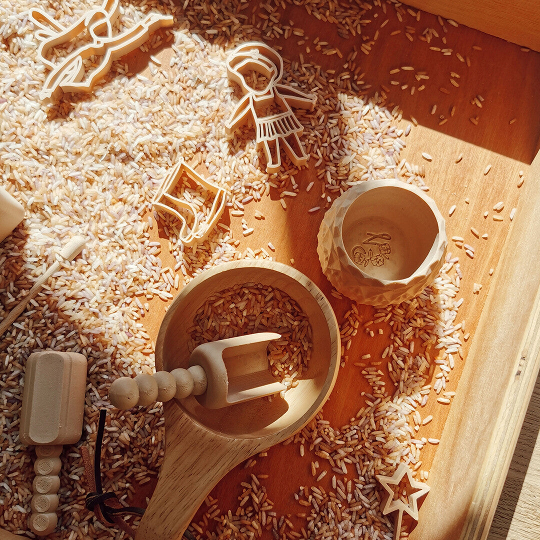 Een beige sensorisch speelgoed schepje ligt in een houten kom, omringd door rijst, uitsteekvormpjes en andere sensorische speelgoedjes.