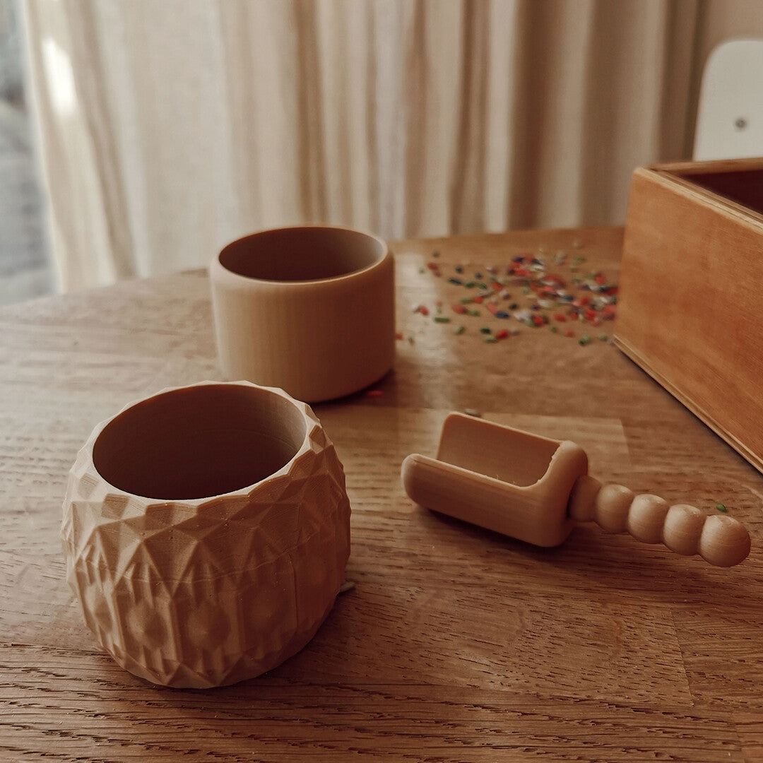 Een grennn beige sensorisch speelgoed schepje ligt op een houten tafel naast twee houten bekers en gekleurde kraaltjes.