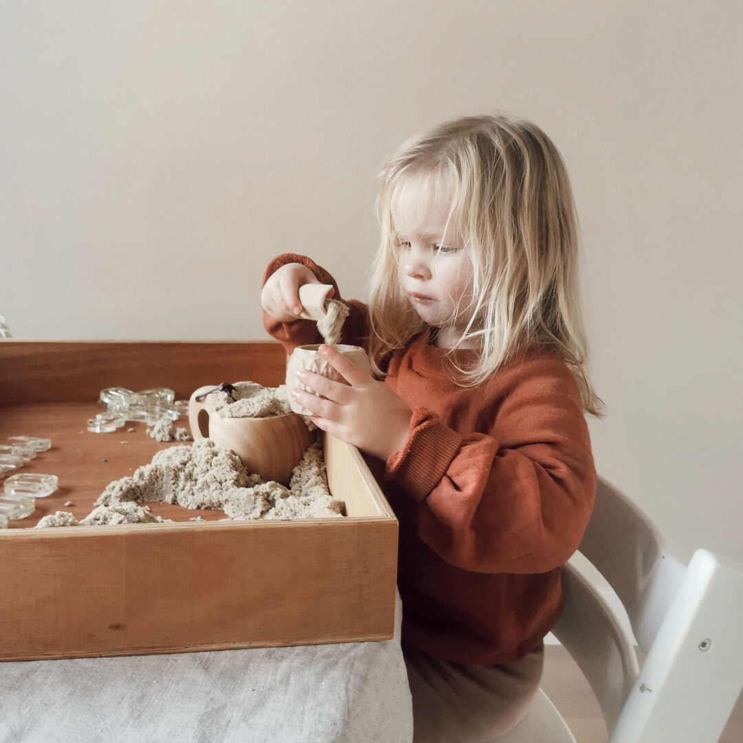 Een kind speelt met zand in een houten speelbak, met een beige speelbakje en een houten schepje.
