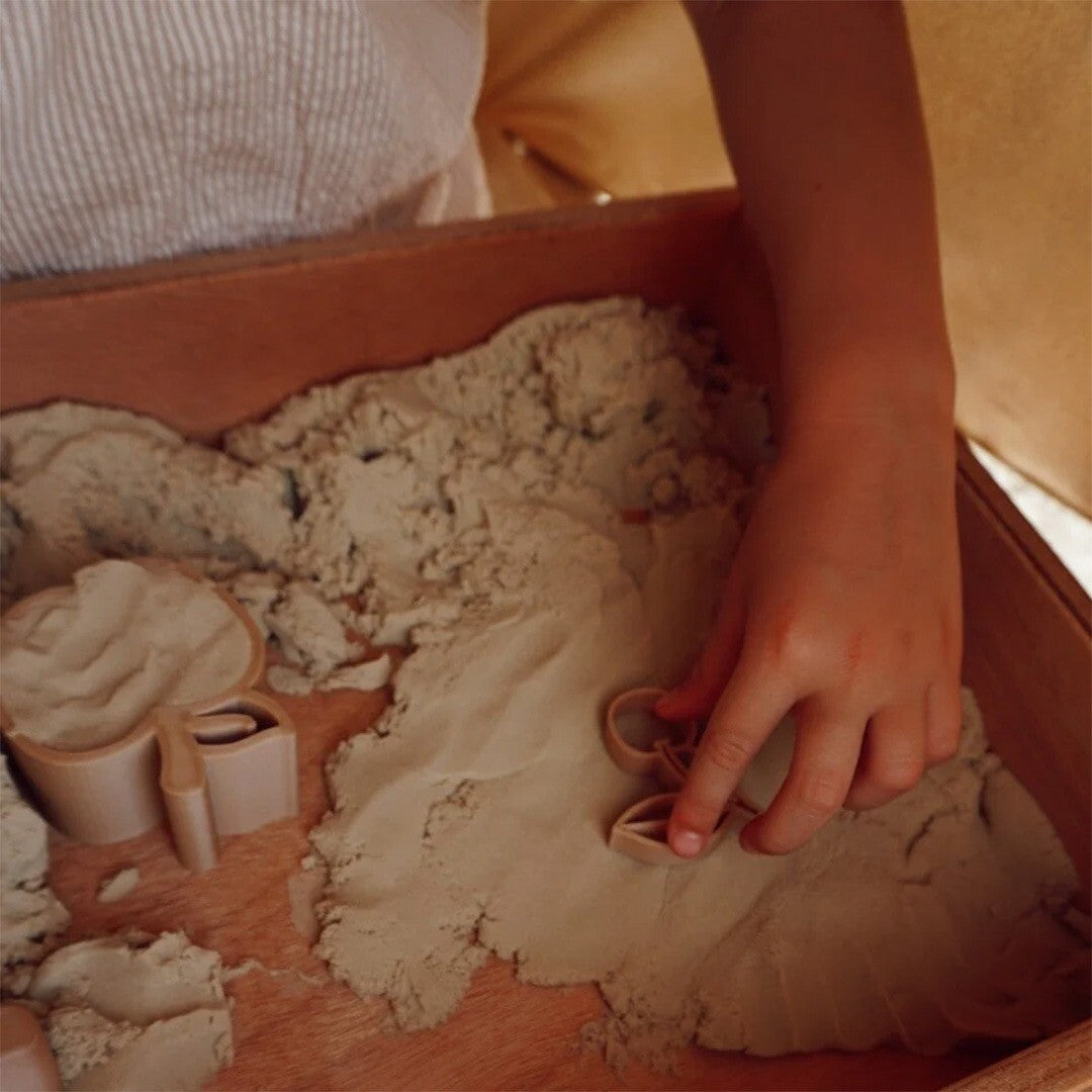 Een kinderhand drukt een kersen uitsteekvorm in het speelzand in een houten zandbak.