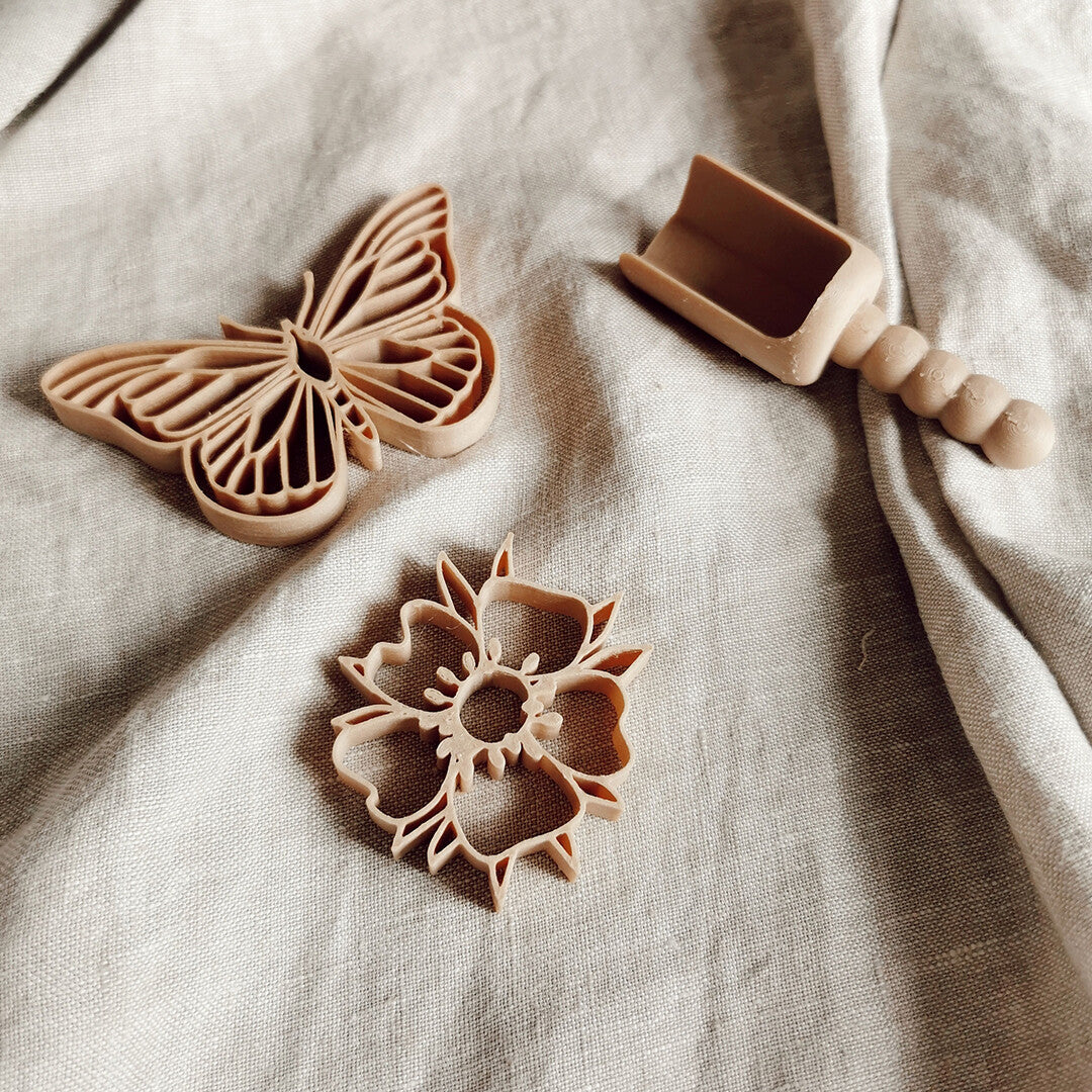 grennn uitsteekvormen voor in de zandbak, een beige vlinder, een bloem en een schepje op een lichte stoffen ondergrond.