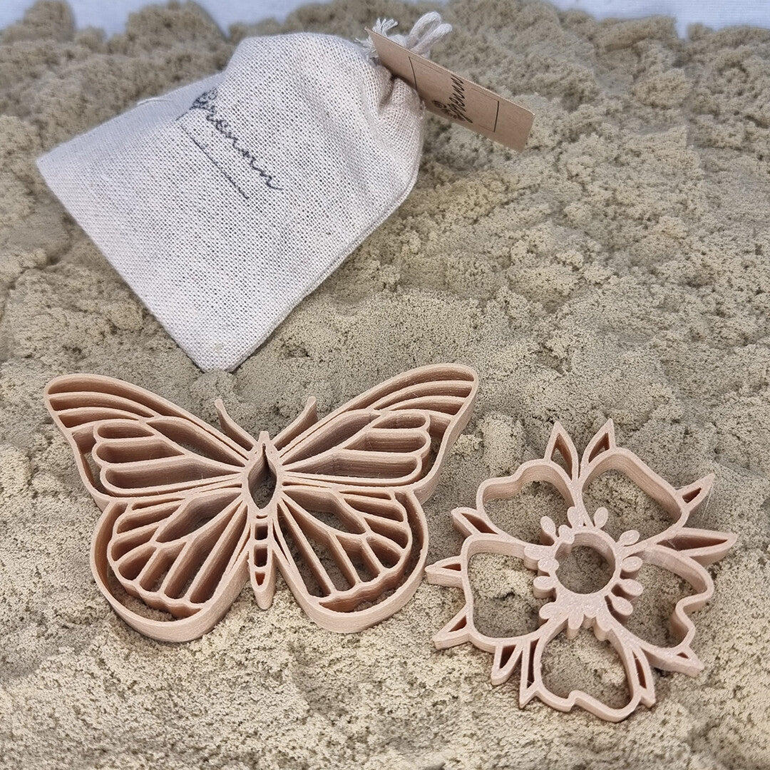 grennn uitsteekvormen voor in de zandbak, een beige vlinder en een bloem op zand, met een linnen zakje op de achtergrond.