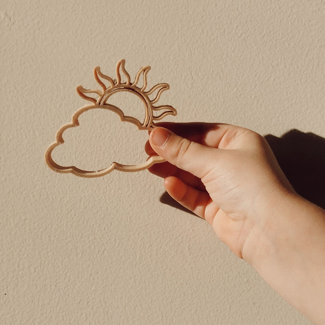 Een kinderhand houdt de grennn zandvorm van een zon met wolk tegen een lichte muur.