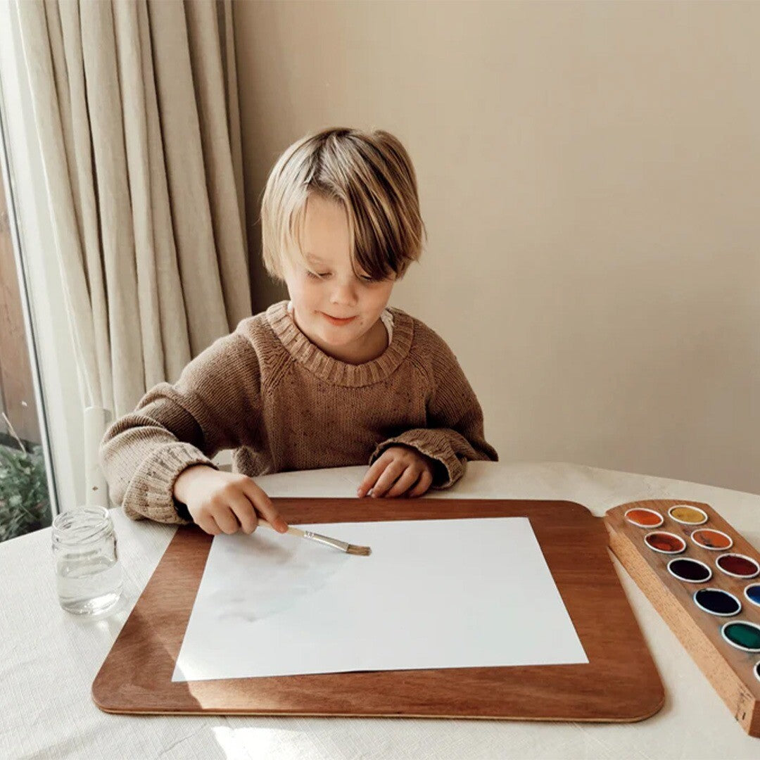 Een kind schildert met waterverf uit de grennn waterverfhouder aan tafel.