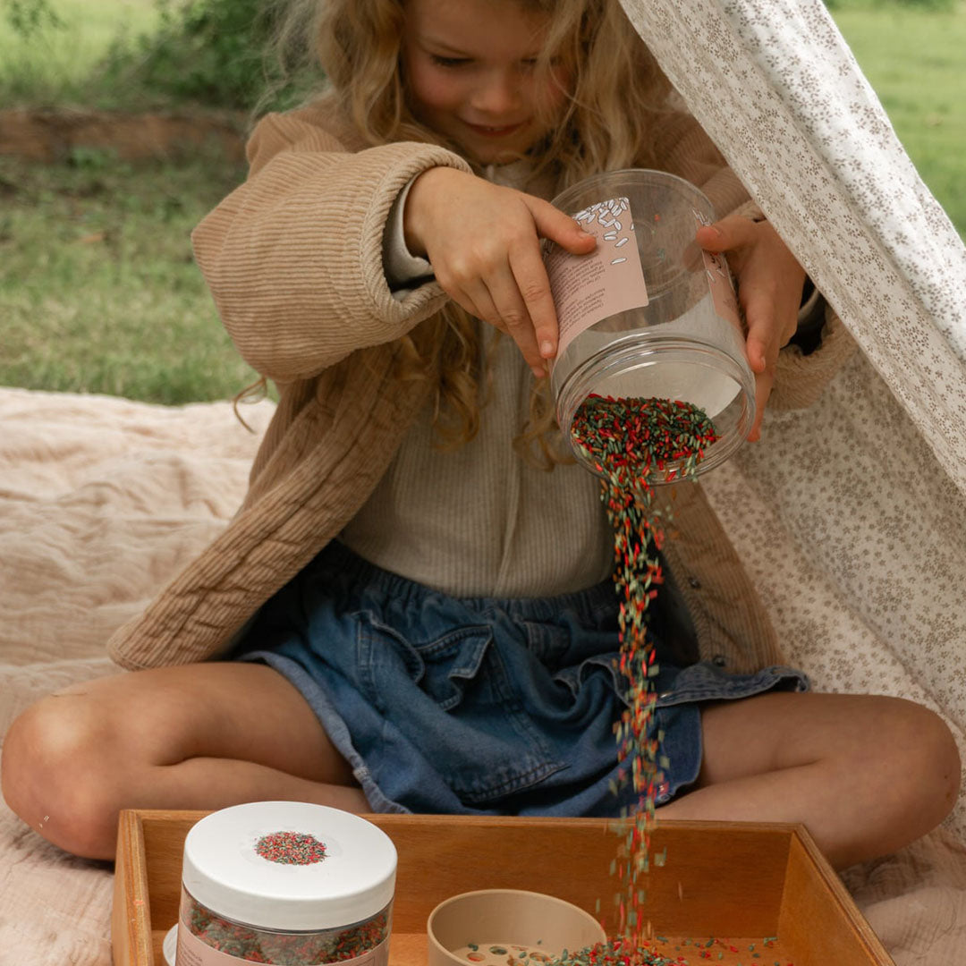 Een kind giet veelkleurige grennn speelrijst uit een pot in een houten speelbak.