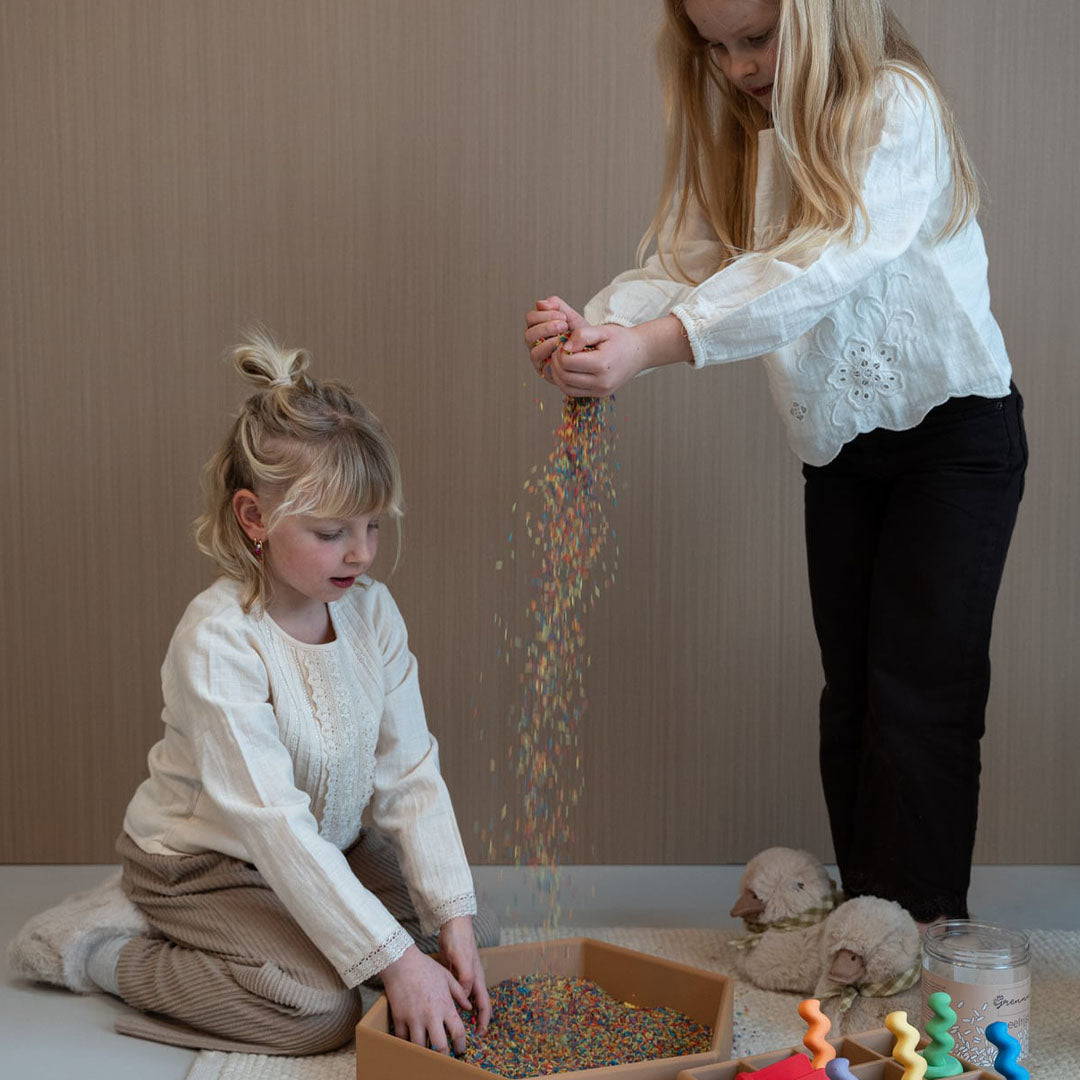 Twee kinderen spelen met veelkleurige speelrijst, waarbij één kind de rijst uitstrooit.