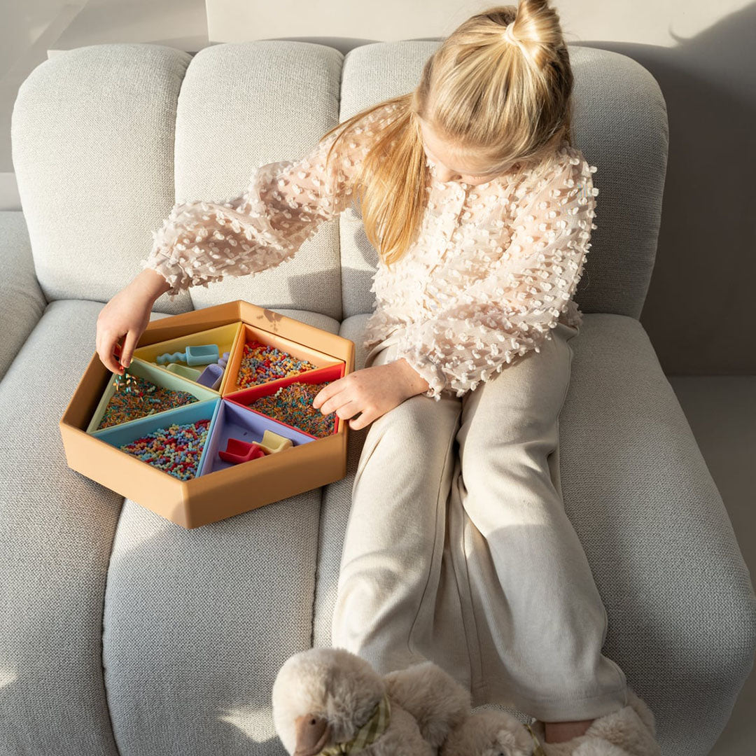 Een kind zit op de bank en speelt met een grote zeshoekige sorteerbak gevuld met grennn speelrijst pastelmix en kleine schepjes.