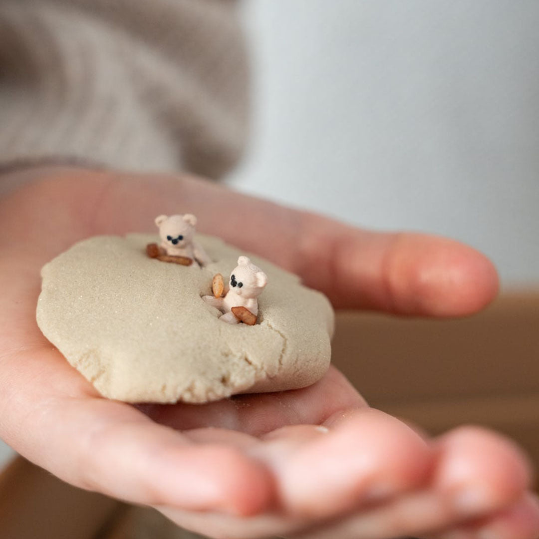 Een hand houdt een bal zand of deeg vast met twee kleine grennn teddybeerfiguurtjes erin.
