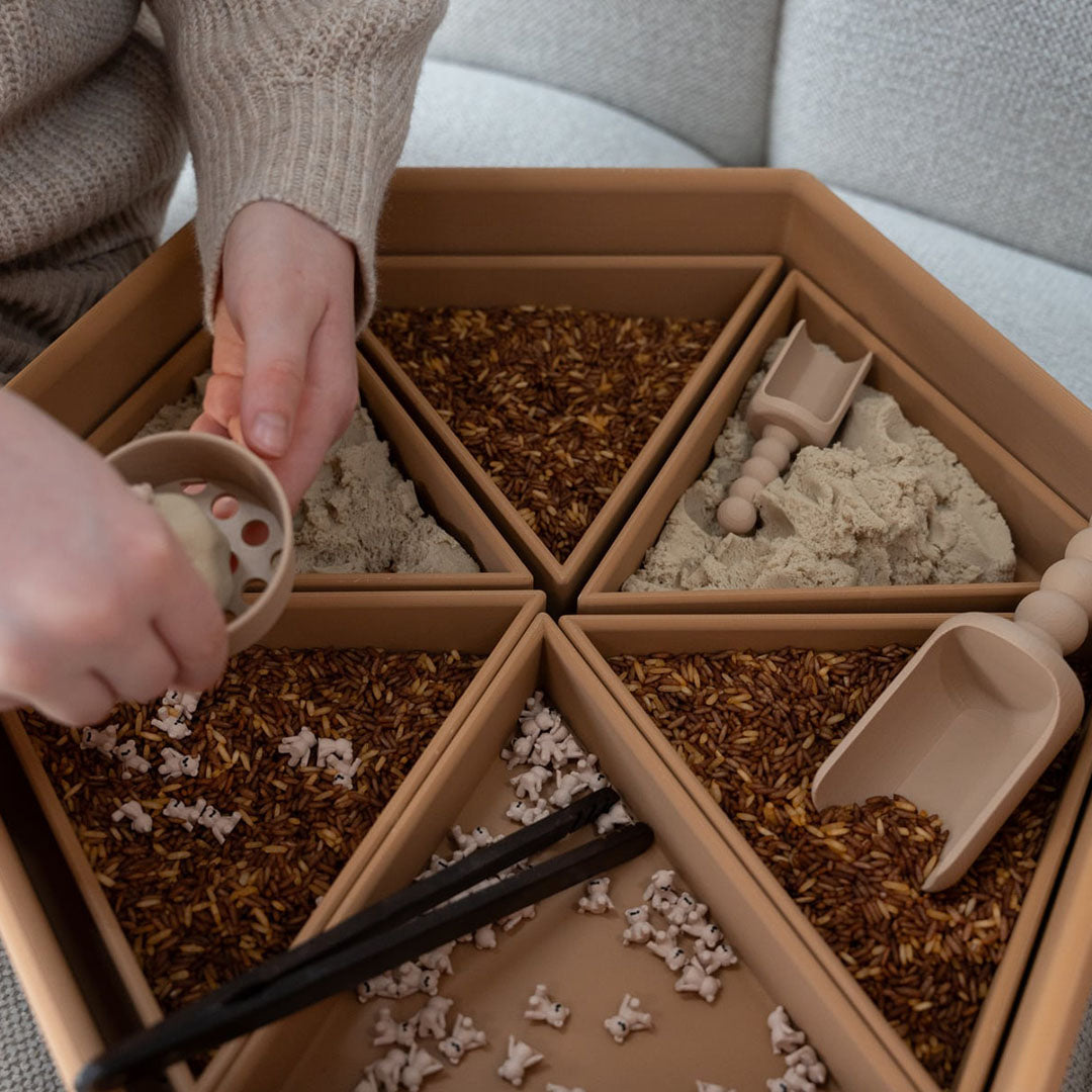 Handen spelen met een grennn sensorische speelbak gevuld met speelrijst, zand en kleine figuurtjes.