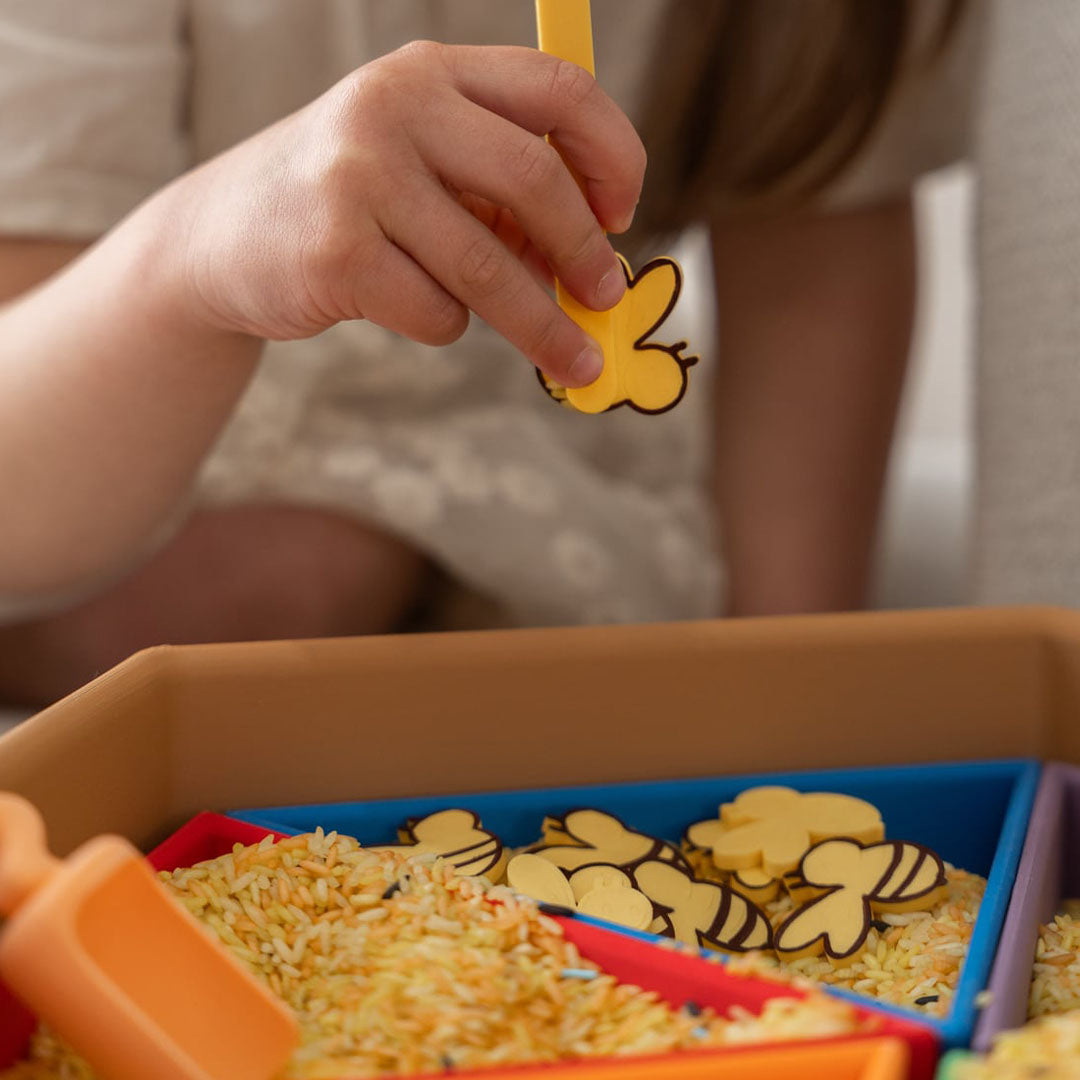 Een kinderhand houdt een geel bijenvormig schepje boven grennn speelrijst met bijenfiguren in een speelbak.