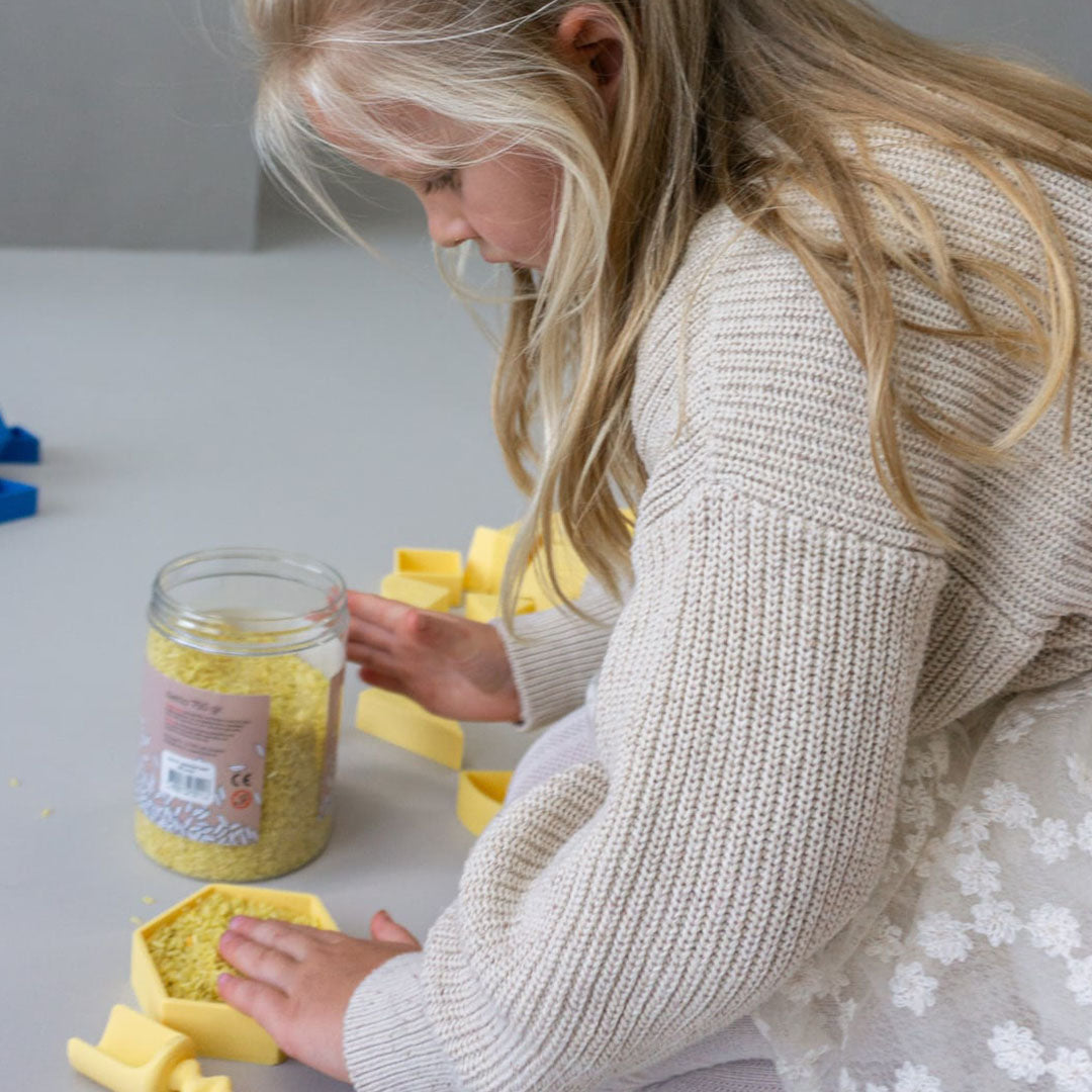 Een kind speelt geconcentreerd met gele speelrijst uit een pot.