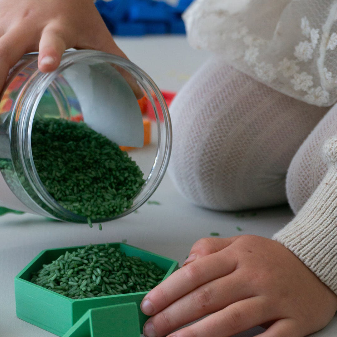 Kinderhanden gieten groene speelrijst uit een pot in een groen bakje.