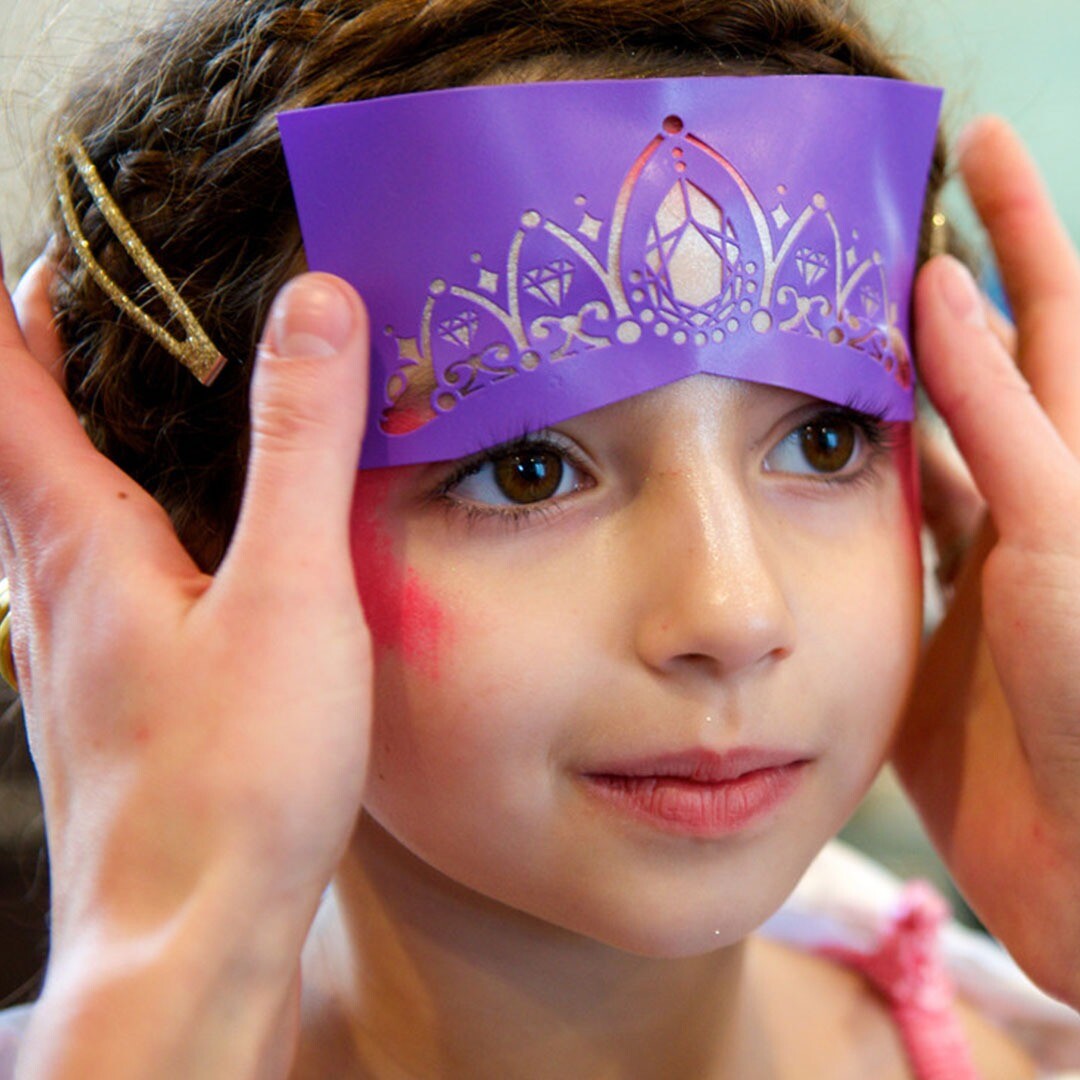 Een meisje met roze schmink op haar wangen draagt een paars prinsessen schminkstencil als tiara op haar voorhoofd.