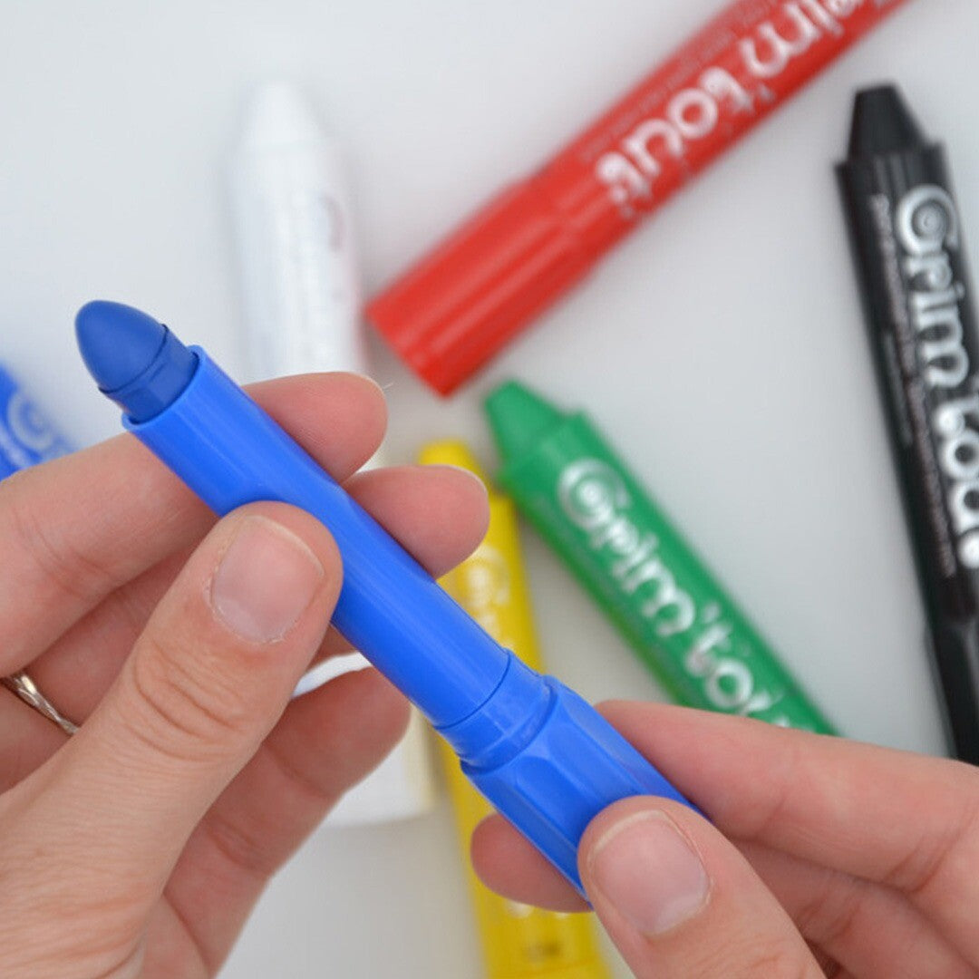 Een hand houdt een blauwe Grim'tout schminkstift vast, met andere kleuren op de achtergrond.