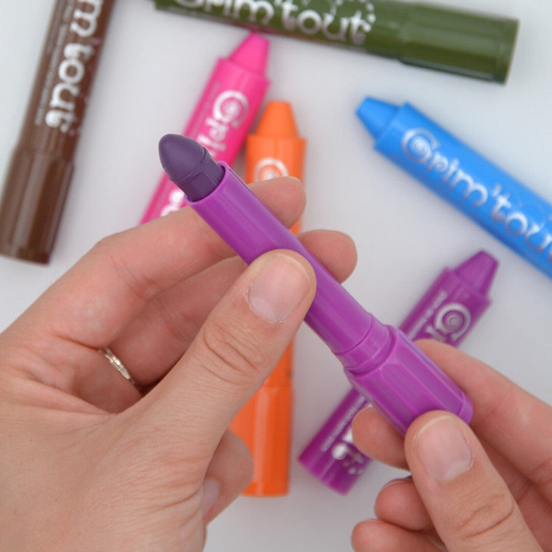 Een hand houdt een paarse Grim'tout schminkstift vast, met andere kleuren op de achtergrond.