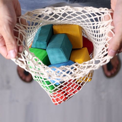 Een persoon houdt een netje vast gevuld met kleurrijke Grimm's abstracte houten bouwblokken.