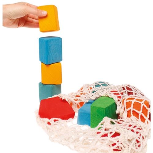 Een hand stapelt kleurrijke Grimm's abstracte houten bouwblokken, met een netje vol blokken ernaast.