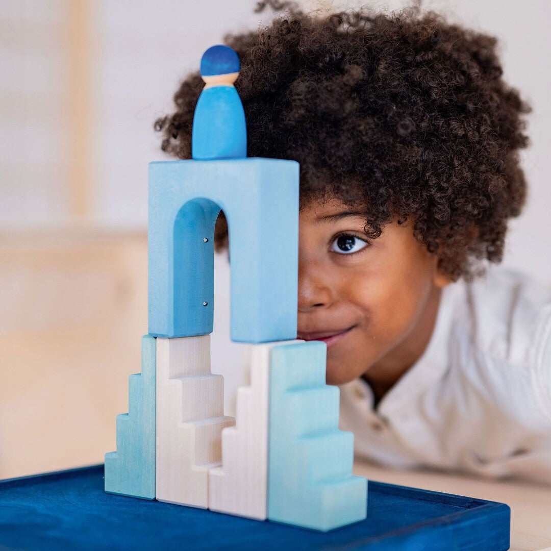 Een kind kijkt lachend achter een bouwwerk van Grimm's houten bouwblokken poollicht vandaan, met een blauw poppetje erop.