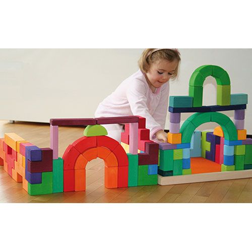 Een kind speelt op een houten vloer met een groot, kleurrijk bouwwerk van Grimm's houten bouwblokken Romaans.