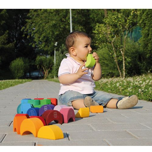 Een baby zit buiten op de grond en bijt op een groen grimm's houten maanhuisje, met andere gekleurde blokken eromheen.