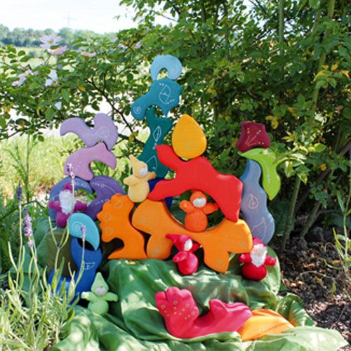 Een collectie Grimm's houten blokken en stoffen figuren, waaronder het oranje Huis Sole, opgesteld in een tuin met bloemen.