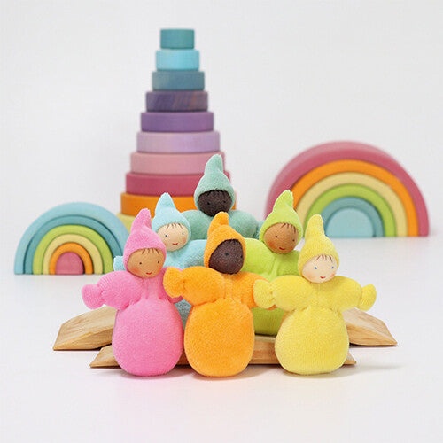 Zes grimm's kabouter poppen in pastelkleuren staan op een houten basis met op de achtergrond twee houten regenboog stapelaars en een toren van houten schijven.