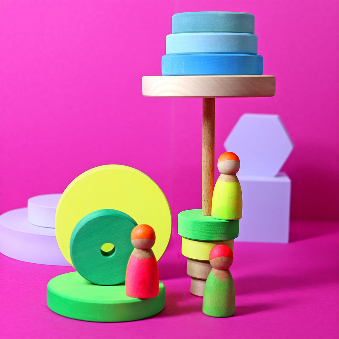 Een collectie Grimm's houten popjes en stapelspeelgoed in diverse felle kleuren op een roze achtergrond.