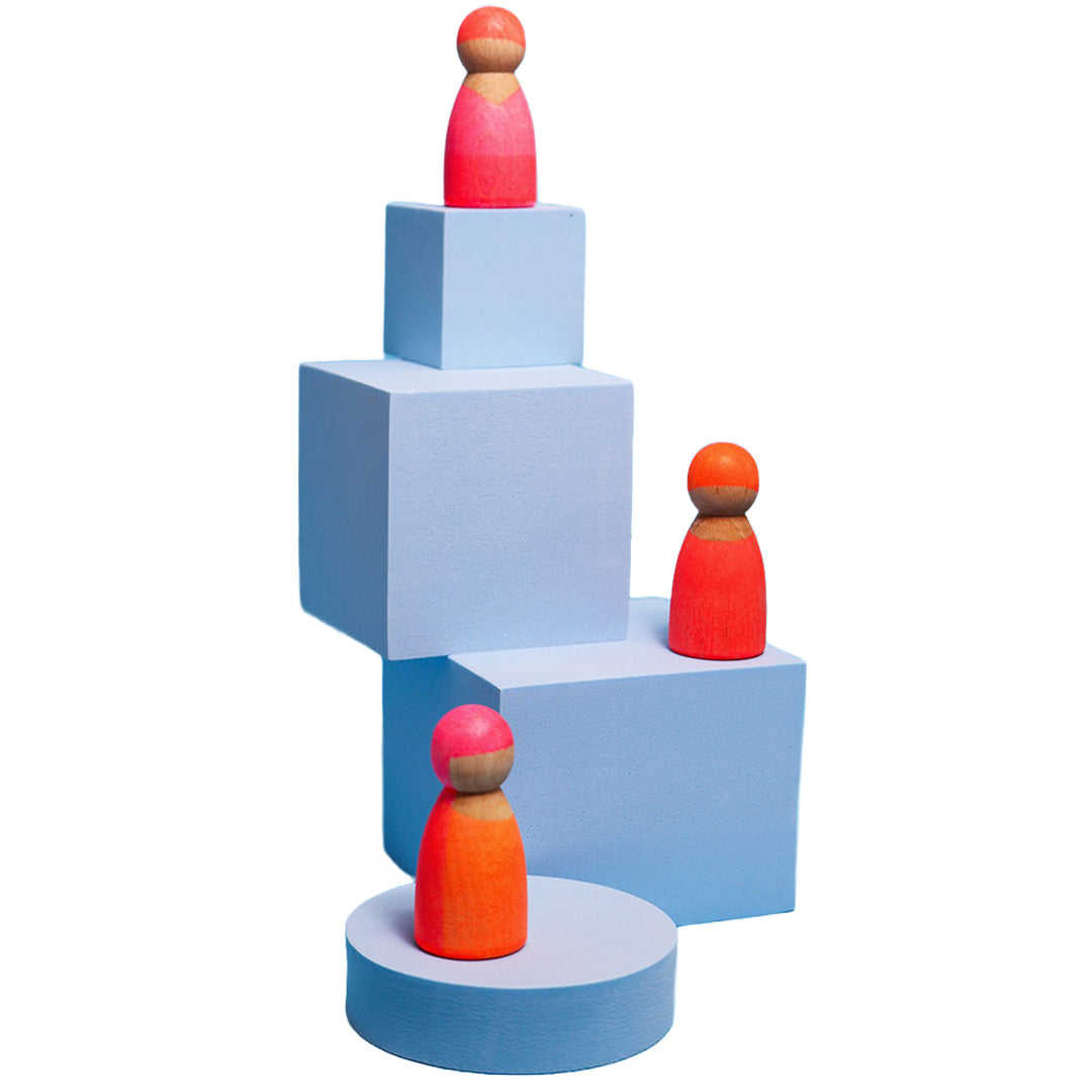 Drie Grimm's neon roze houten popjes staan op lichtblauwe blokken.