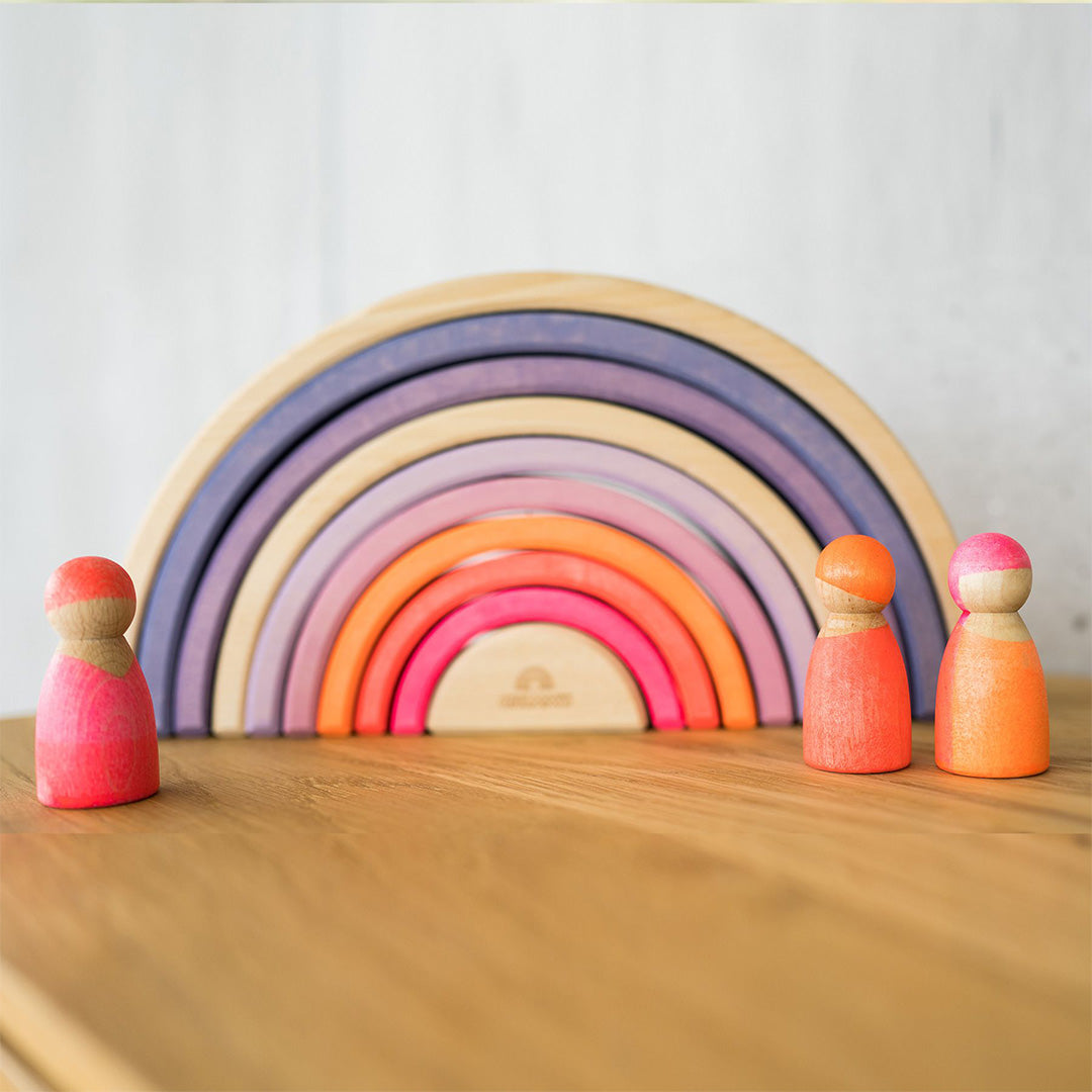 Twee Grimm's neon roze houten popjes staan voor een houten regenboogstapelaar.