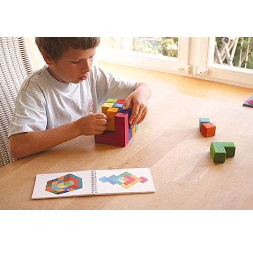 Een kind bouwt een kleurrijke Grimm's geometrische puzzel op een houten tafel, met een open voorbeeldboekje ernaast.