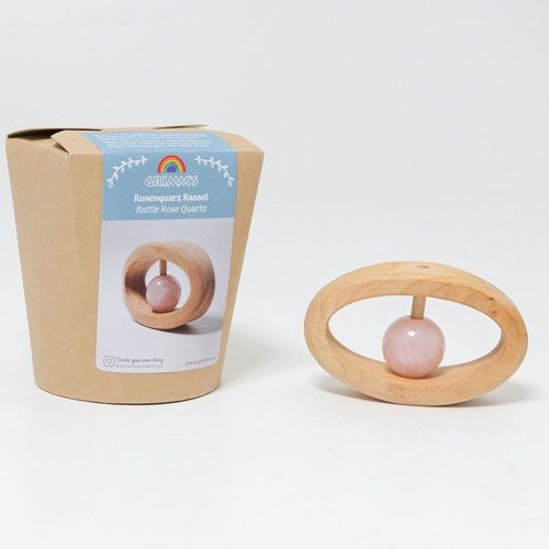 Grimm's houten rammelaar-bijtring met rozenkwarts naast de productverpakking.