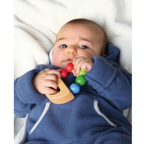 Een baby in een blauw pakje ligt op een witte deken en speelt met de Grimm's regenboog rammelaar.