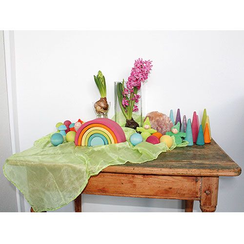 Een houten tafel versierd met Grimm's houten speelgoed in pastelkleuren, waaronder een regenboog en kegels, naast roze hyacinten.