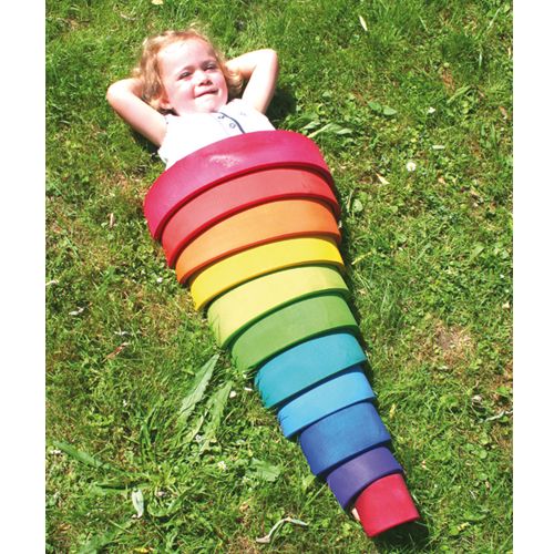 Een lachend kind ligt in het gras, omringd door de grimm's houten regenboog stapeltoren die als een kleurrijke cocon is opgesteld.