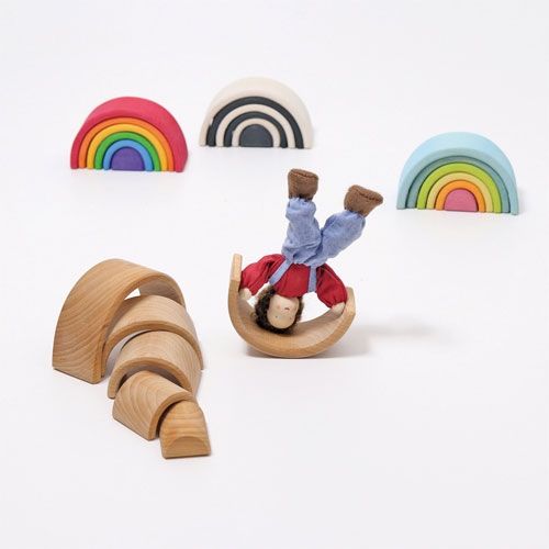 Een collectie Grimm's houten regenboog stapelbogen in verschillende kleuren, met een pop die balanceert op een naturelkleurige boog.