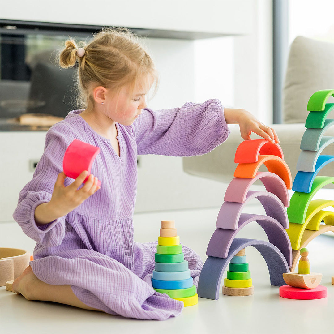 Een meisje speelt met een Grimm's houten regenboog stapeltoren en ander houten stapelspeelgoed.
