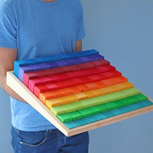 Een persoon houdt een houten bak met Grimm's regenboogkleurige bouwblokken vast.
