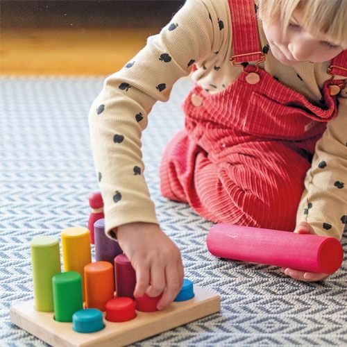 Een kind speelt met de grimm's houten regenboog stapelrollers, waarbij een rode roller in de houten basis wordt geplaatst.
