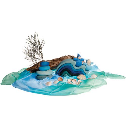 Een Grimm's houten speelset met blauwe golven, gestapelde kiezelstenen en figuren, omringd door schelpen en zijden doeken die water voorstellen.
