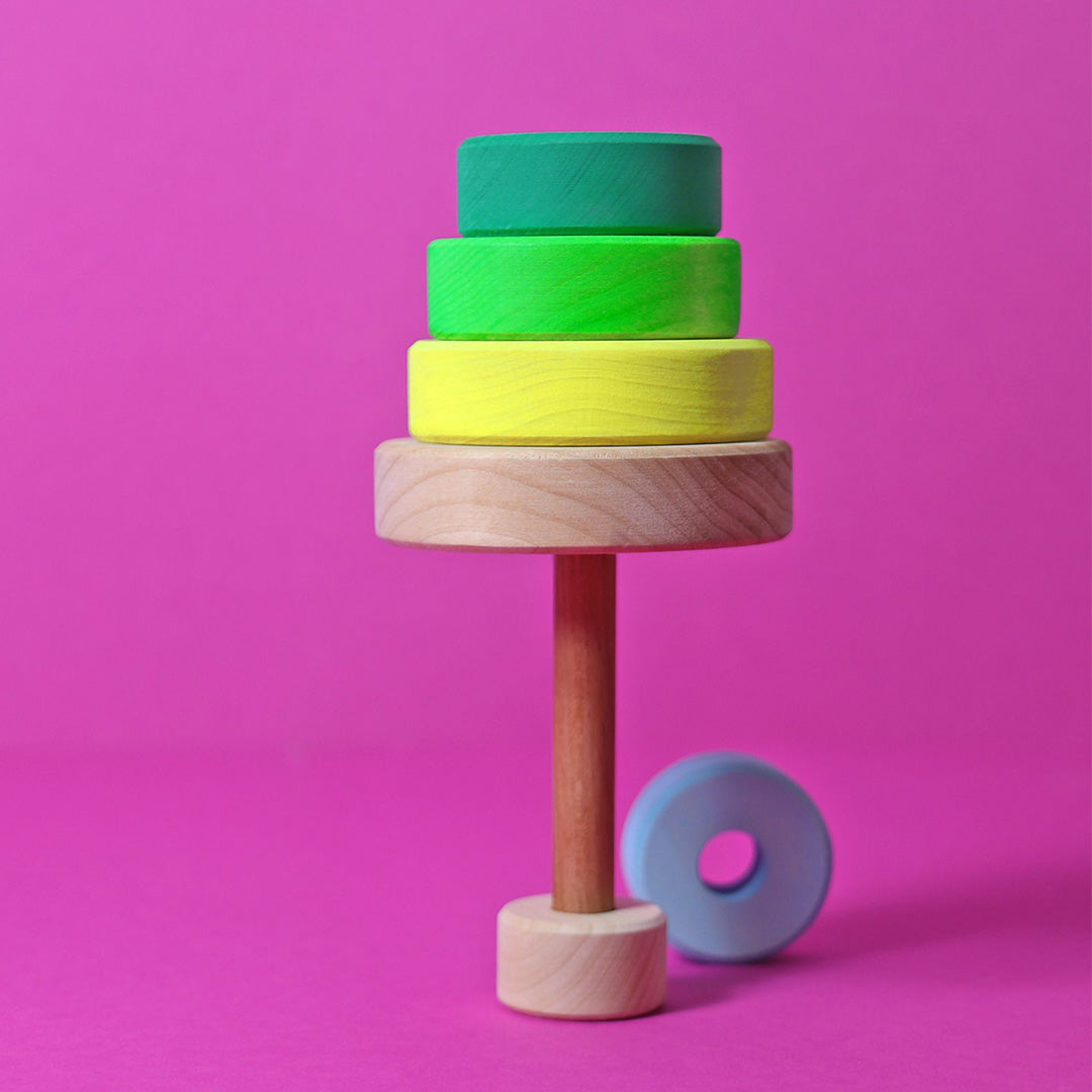 Grimm's houten stapelspeelgoed met neon groene en gele ringen op een roze achtergrond, met een losse blauwe ring ernaast.