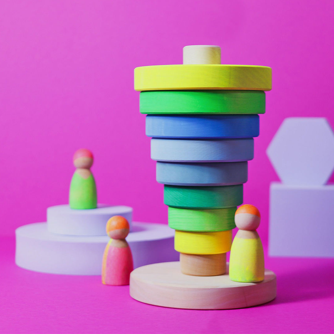 Een Grimm's houten stapeltoren met veelkleurige ringen en twee houten poppetjes op paarse platforms, op een roze achtergrond.