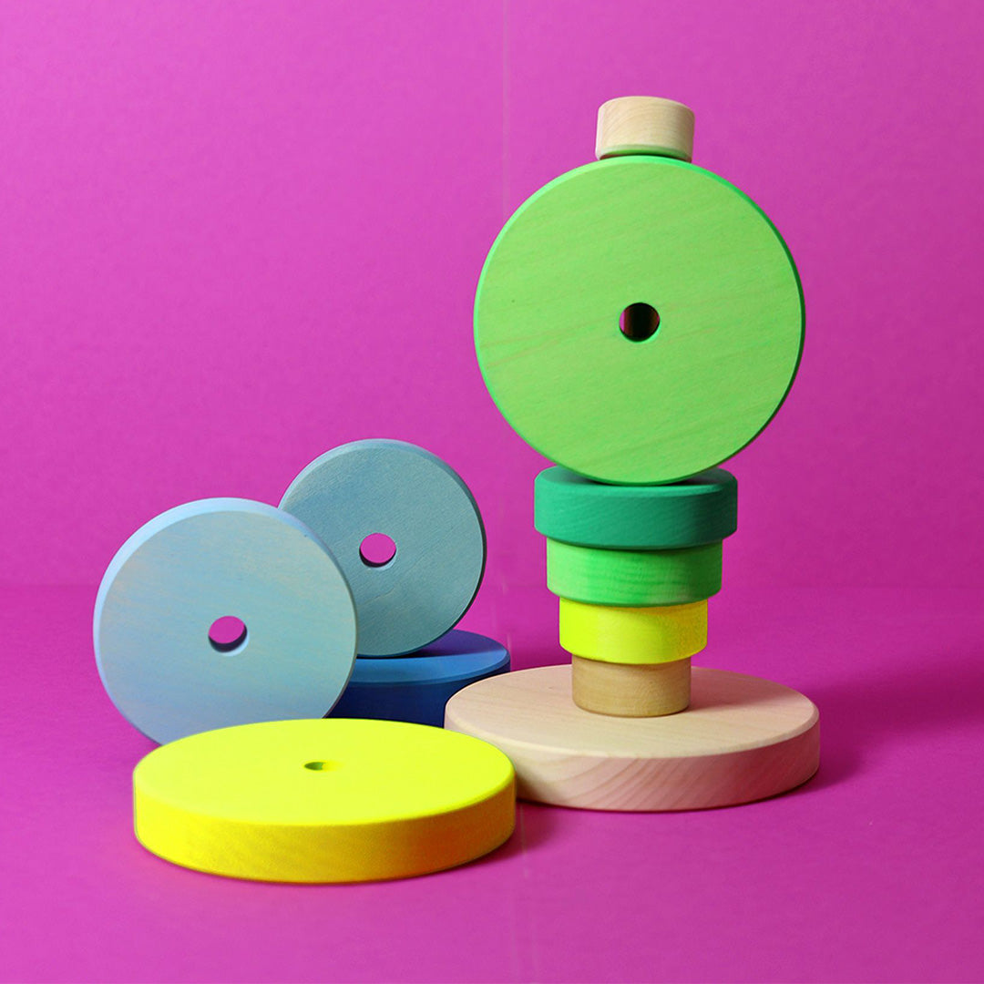 Een Grimm's houten stapeltoren met groene en gele ringen, en losse blauwe en gele ringen ernaast, op een roze achtergrond.