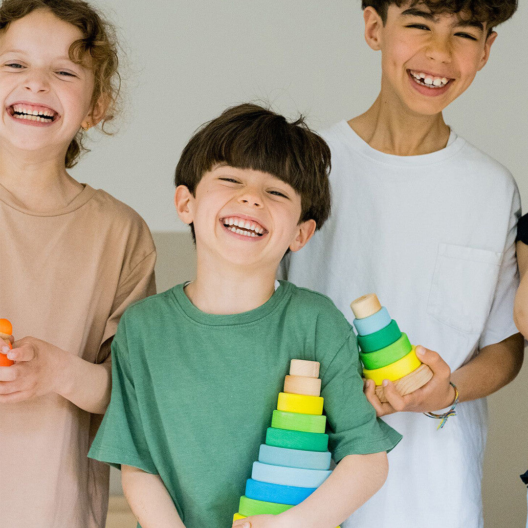 Drie lachende kinderen, waarvan één een Grimm's houten stapeltoren voor baby's en peuters vasthoudt.