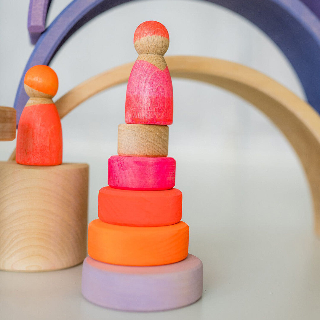 Grimm's houten stapeltoren voor baby's met neonkleurige ringen staat naast twee houten poppetjes en een regenboogboog.