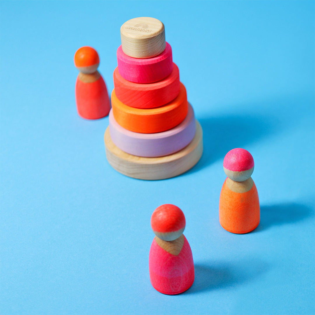 Grimm's houten stapeltoren voor baby's met neonkleurige ringen staat op een blauwe achtergrond, omringd door drie houten poppetjes.